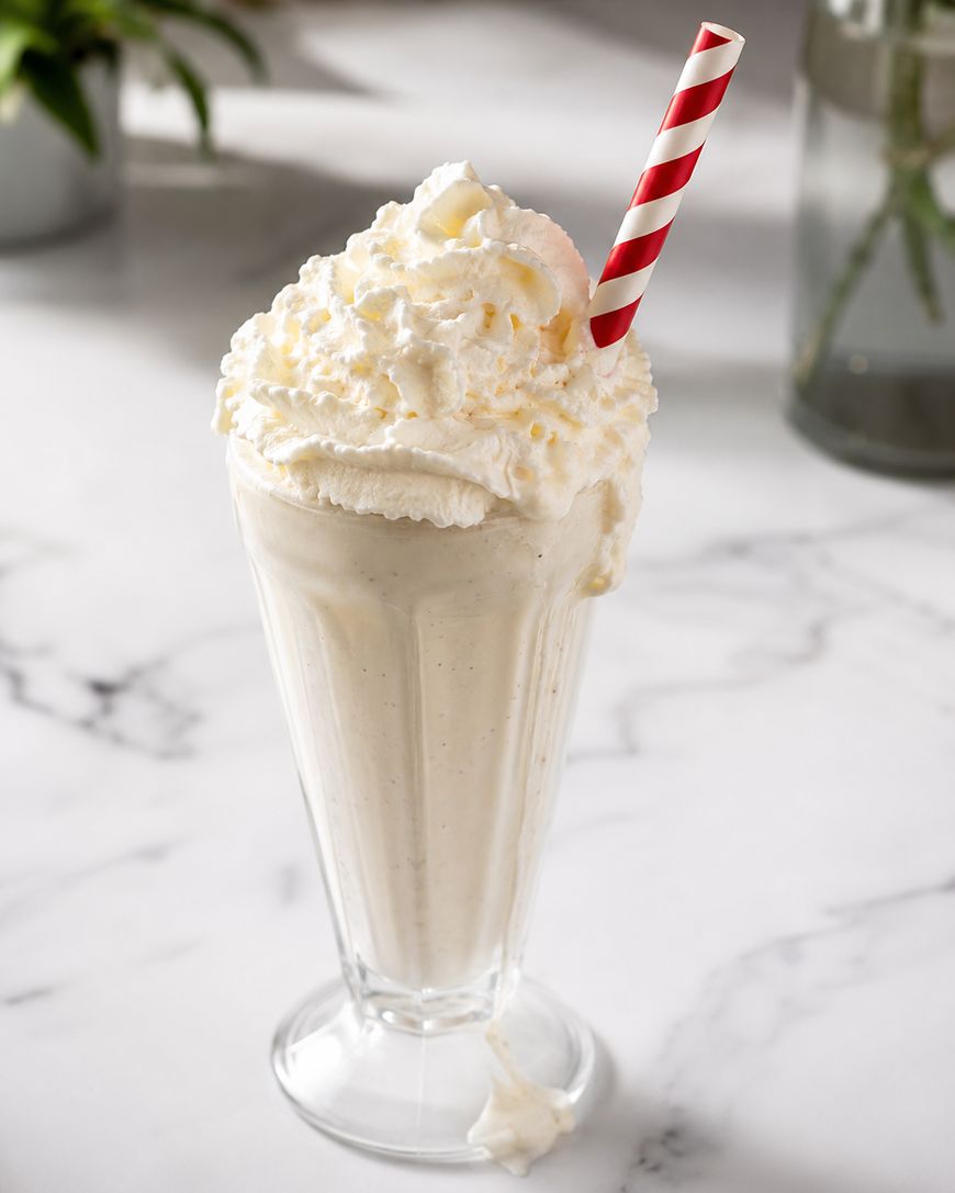 A tall glass filled with a vanilla milkshake, topped with whipped cream and a red and white striped straw, sits on a white marble surface. A small dollop of whipped cream is on the base of the glass.