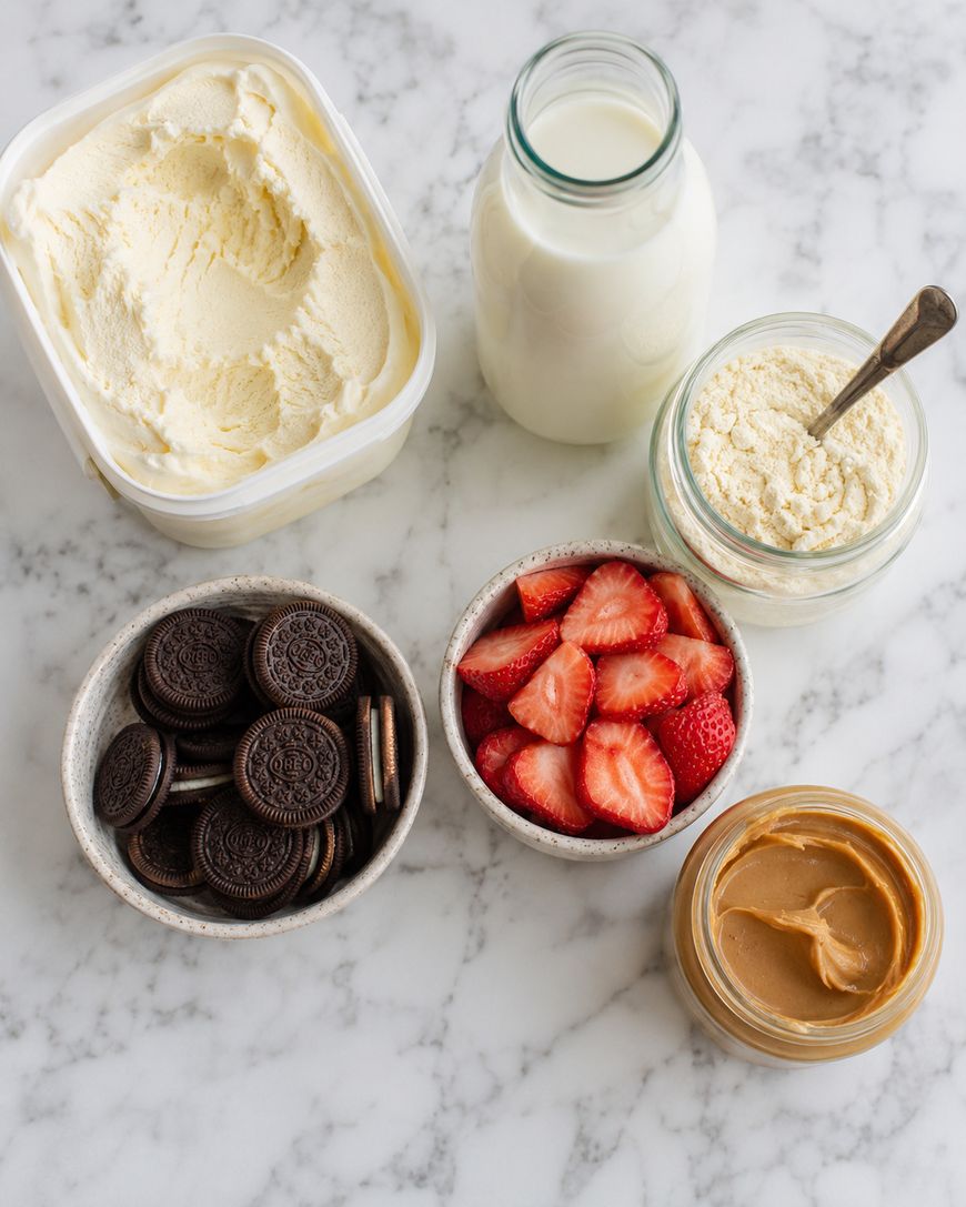 A marble surface displays containers of vanilla ice cream, milk, protein powder, chocolate sandwich cookies, sliced strawberries, and creamy peanut butter.