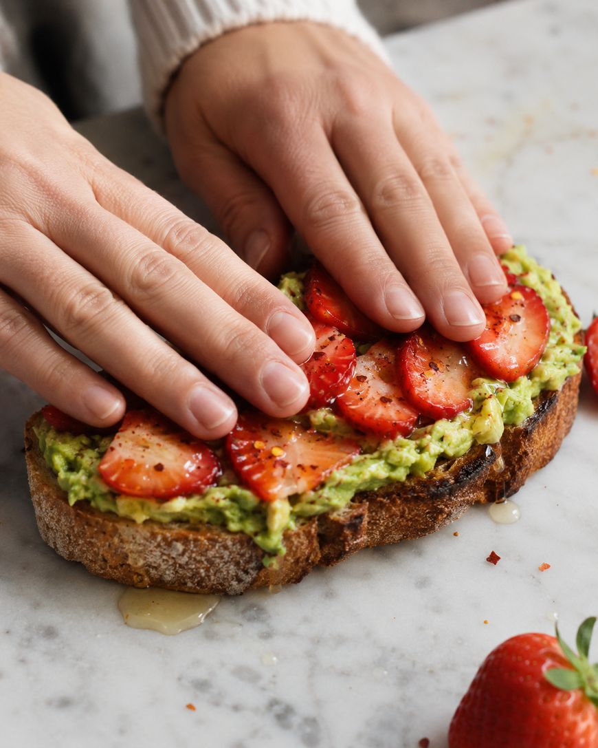 Two hands press gently on a slice of toast topped with mashed avocado, sliced strawberries, and a drizzle of honey, on a marble surface with whole strawberries nearby.