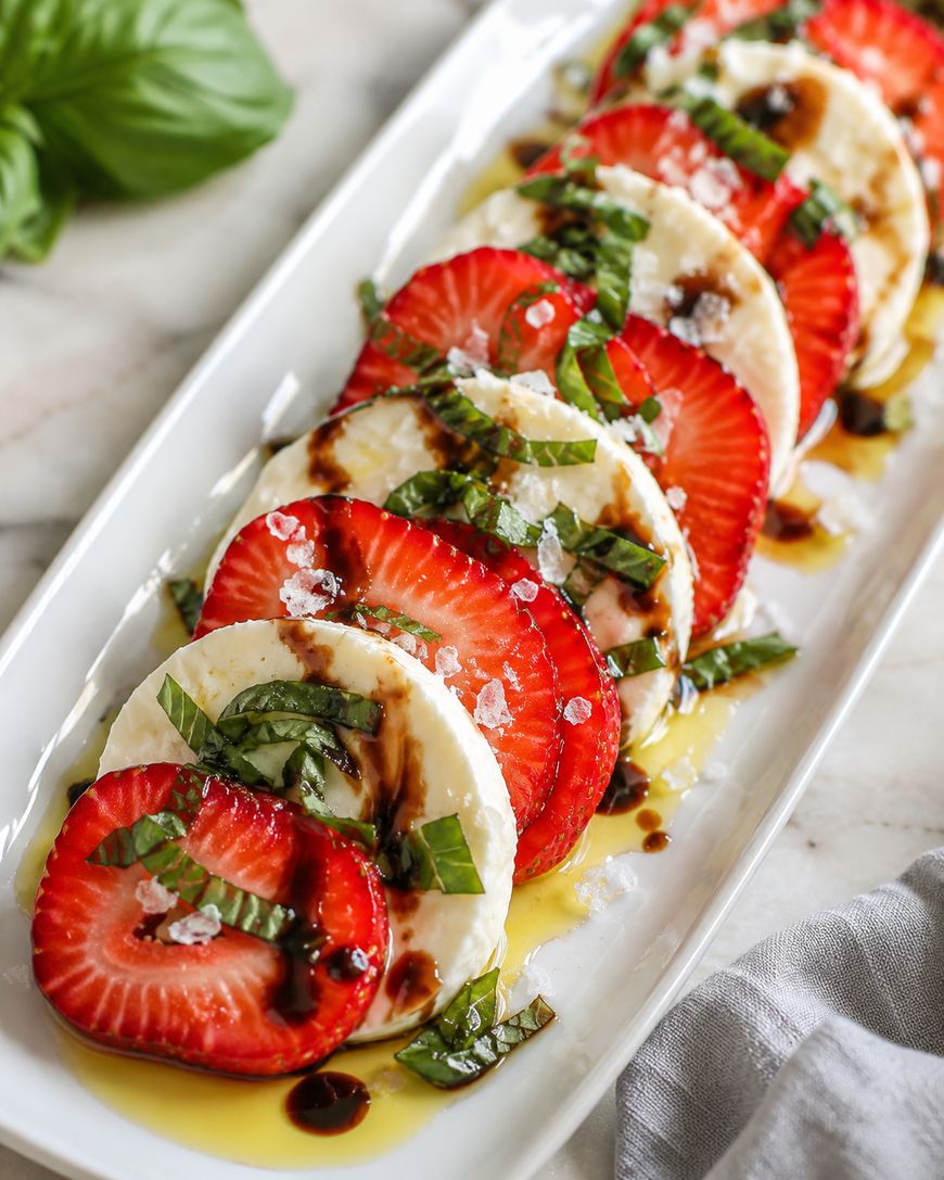 A rectangular white plate with alternating slices of fresh mozzarella and strawberries, topped with chopped basil, sea salt, olive oil, and a drizzle of balsamic glaze.