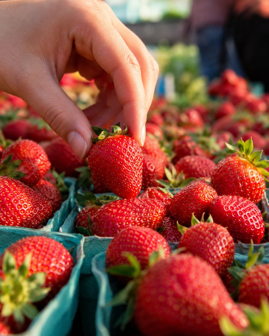 A close-up of a hand picking a ripe strawberry from a green carton filled with fresh strawberries at an outdoor market.