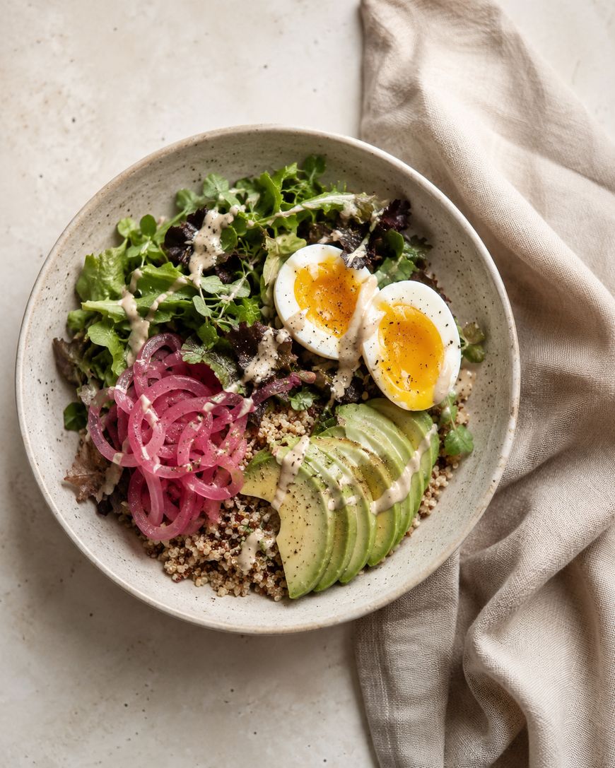 A bowl with greens, sliced avocado, pickled red onions, quinoa, and a halved soft-boiled egg, drizzled with creamy dressing, sits on a light surface next to a beige cloth napkin.