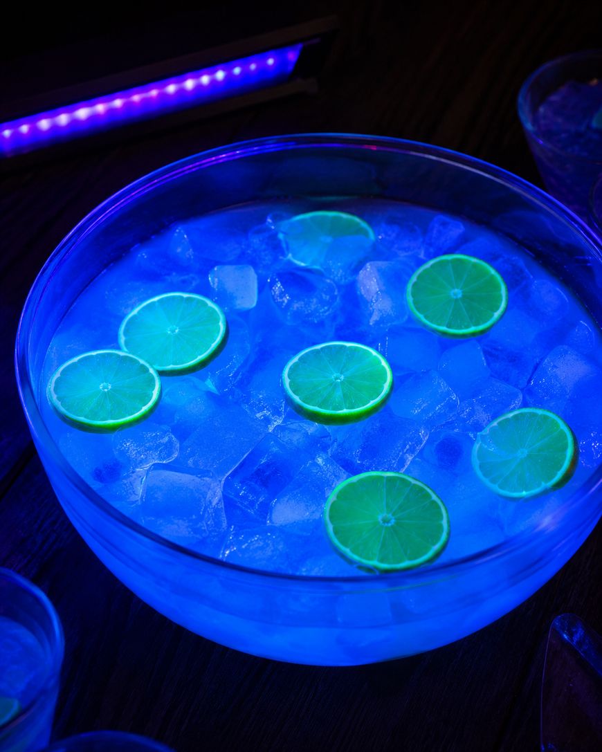 A bowl filled with ice cubes and sliced limes glows blue under blacklight, creating a vibrant, neon effect. The drink is surrounded by darkness, highlighting the bowls bright, fluorescent appearance.