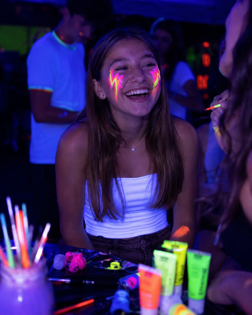 A smiling girl with neon face paint in the shape of lightning bolts enjoys a party under blacklights, surrounded by glowing paint tubes and brushes. Other people with neon designs are in the background.
