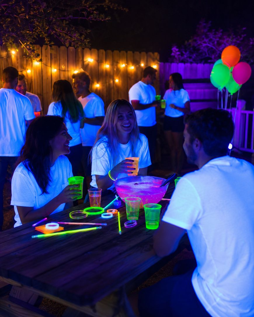 A group of people wearing white shirts and holding neon cups and glow sticks enjoy a nighttime outdoor party at a picnic table, with string lights and balloons in the background.