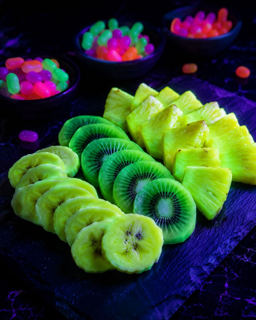 Sliced golden kiwi, green kiwi, and pineapple arranged on a black serving board, with bowls of colorful neon jelly candies in the background. The scene is brightly lit with vibrant colors.