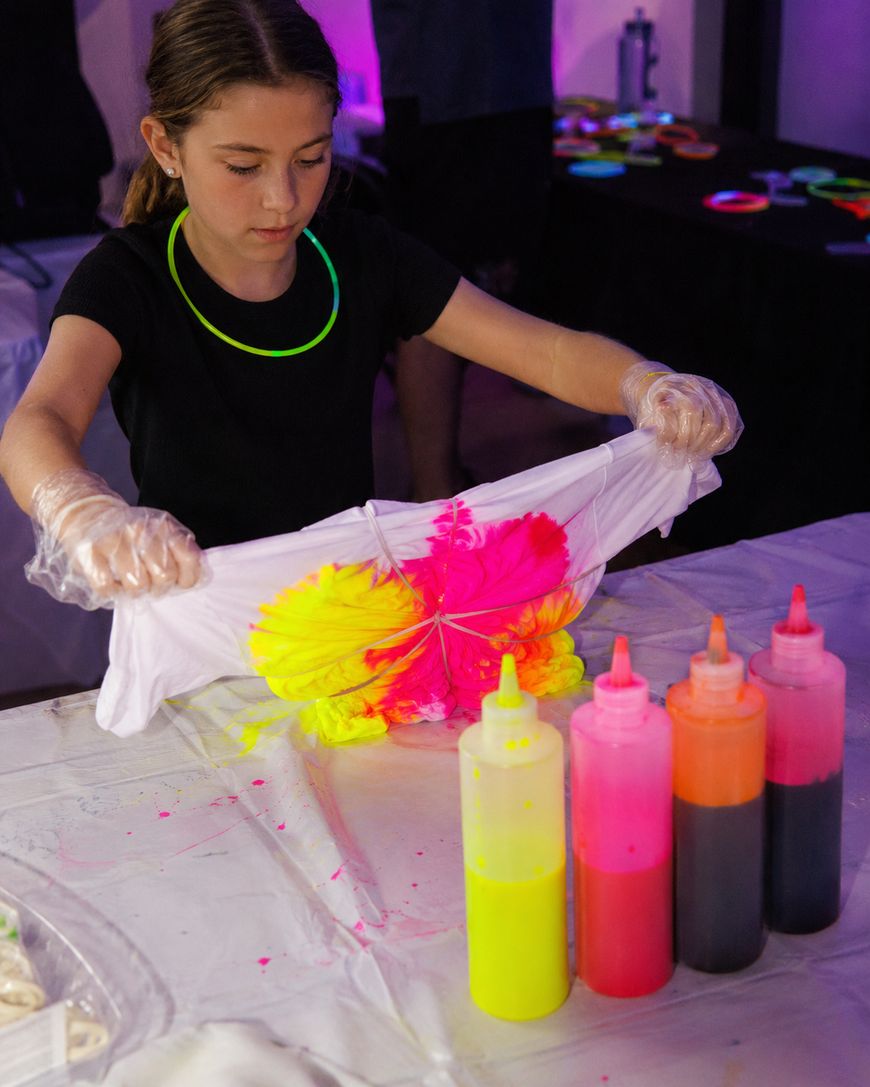 A girl wearing gloves and a black shirt is folding a white T-shirt with bright yellow and pink dye on it. Bottles of colorful dye are lined up on the table in front of her.