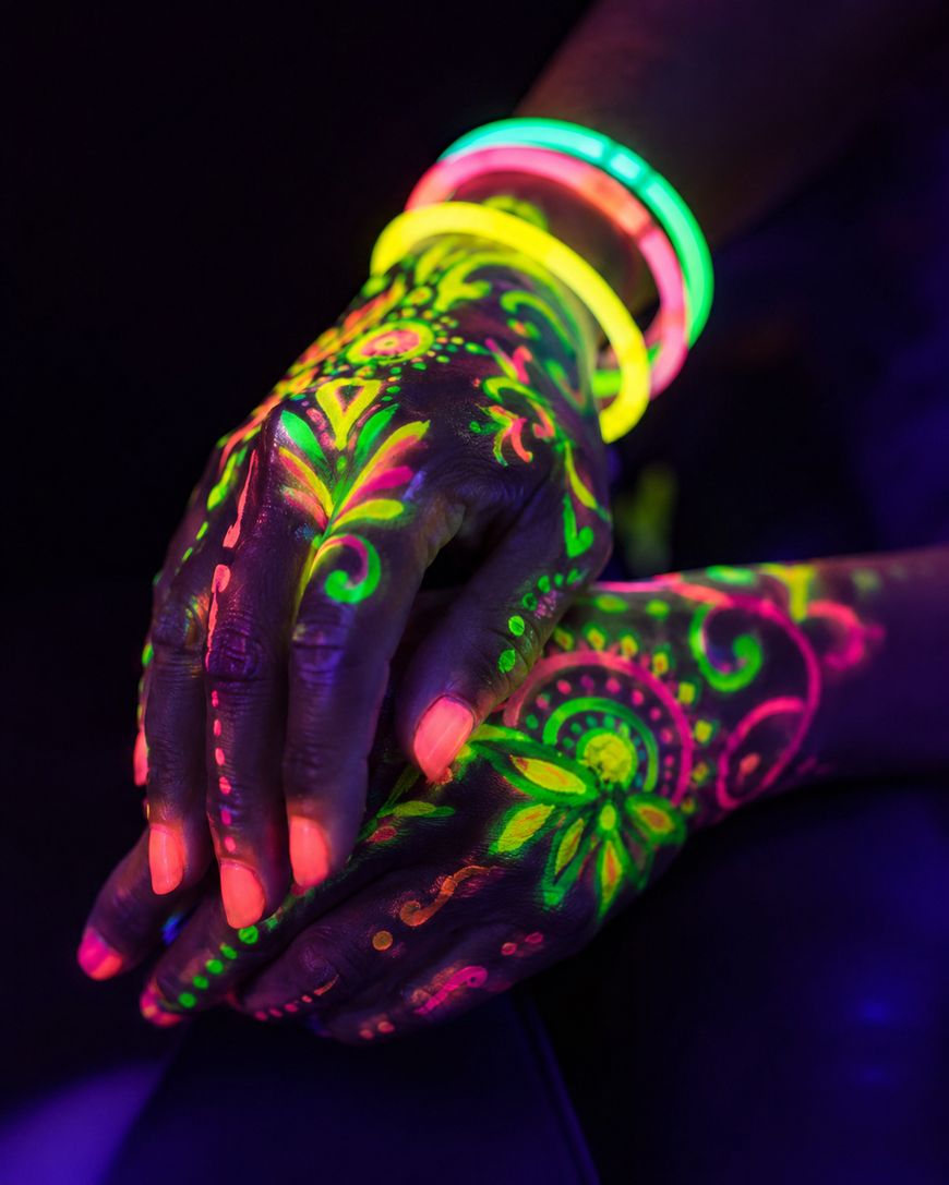 Hands decorated with colorful neon henna designs glow under blacklight. The person is also wearing several glowing bracelets in yellow, green, and pink, creating a vibrant, fluorescent effect.