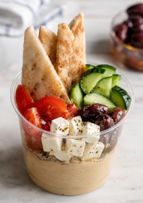 A clear plastic grazing cup filled with hummus, topped with pita bread slices, cherry tomatoes, cucumber slices, kalamata olives, and cubed feta cheese—one of the best grazing cup ideas for easy and stylish party appetizers.