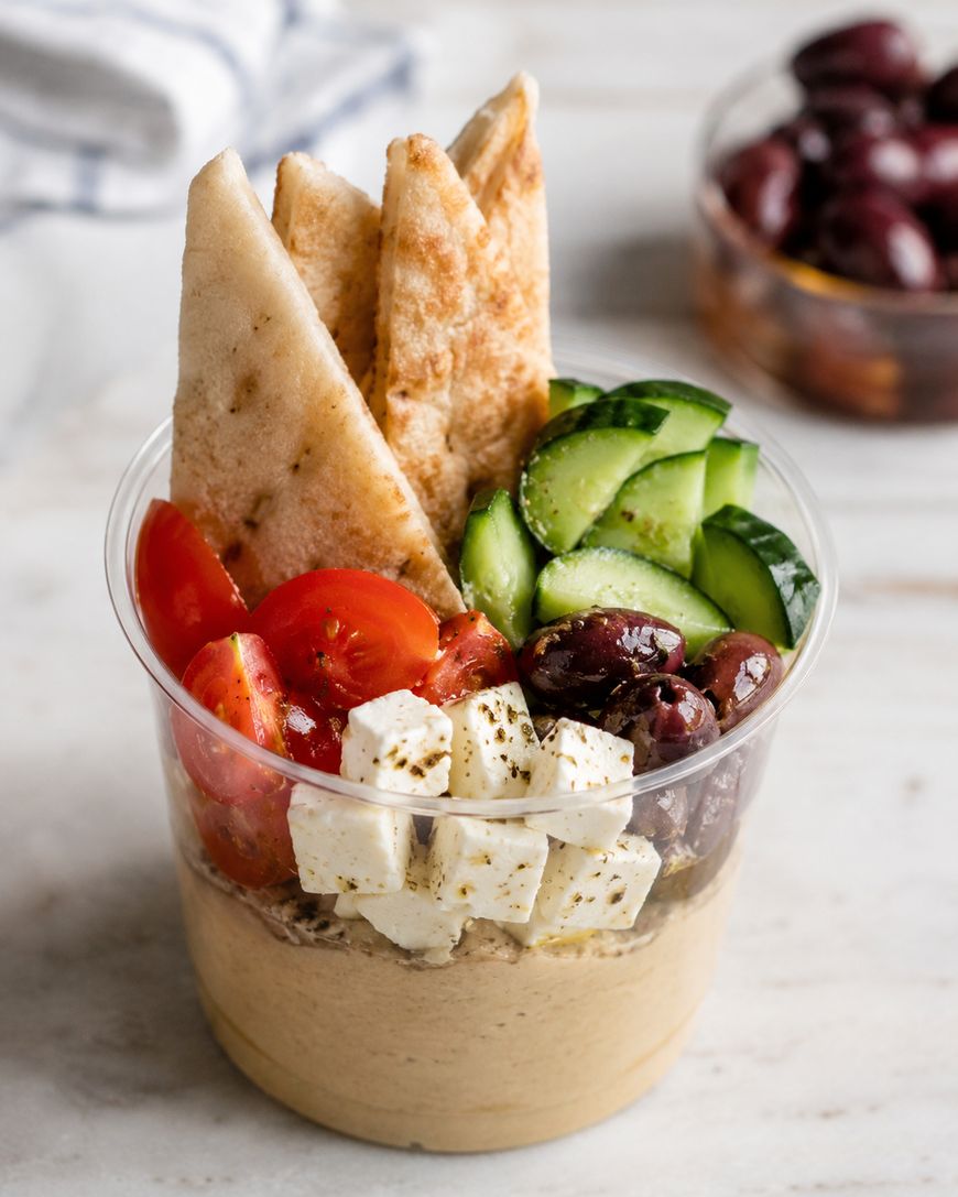 A clear plastic cup filled with hummus, topped with pita bread slices, cherry tomato halves, cucumber slices, Kalamata olives, and cubes of feta cheese. A bowl of more olives is in the background.