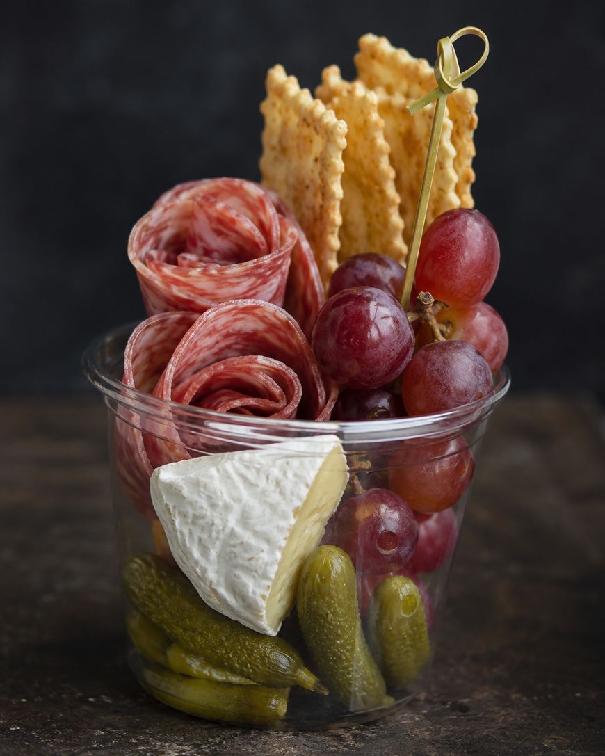 A clear plastic cup filled with rolled salami, red grapes on a skewer, rectangular crackers, a wedge of brie cheese, and several small pickles, arranged neatly against a dark background.