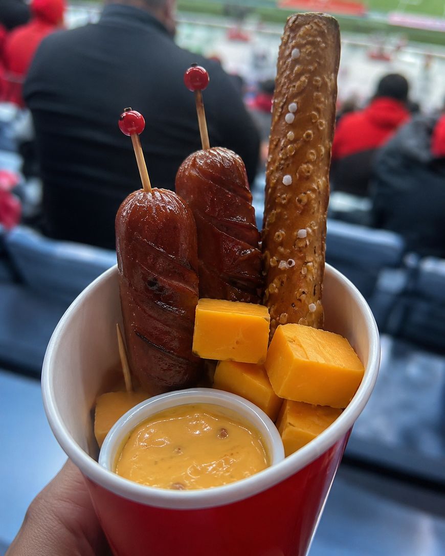 A hand holds a cup filled with two skewered sausages, a soft pretzel stick, cheddar cheese cubes, and a container of cheese dip, with stadium seats and people in the background.