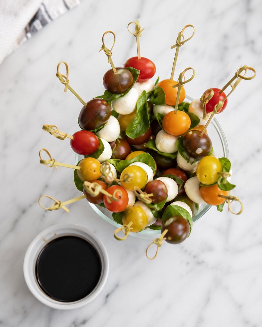 A glass bowl filled with skewers of cherry tomatoes, mozzarella balls, and basil leaves, next to a small ramekin of dark balsamic vinegar, on a white marble surface.