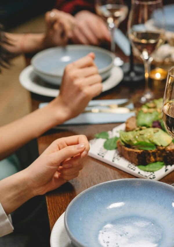 A close-up of a dining table set with blue plates, wine glasses, and a dish topped with avocado. Several people’s hands are visible, suggesting they are gathered for a meal together.