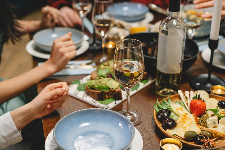 A close-up of a dinner table set with blue plates, wine glasses, a bottle of white wine, candles, and assorted foods including bread, cheese, fruits, and greens. Several people are seated, reaching for food.