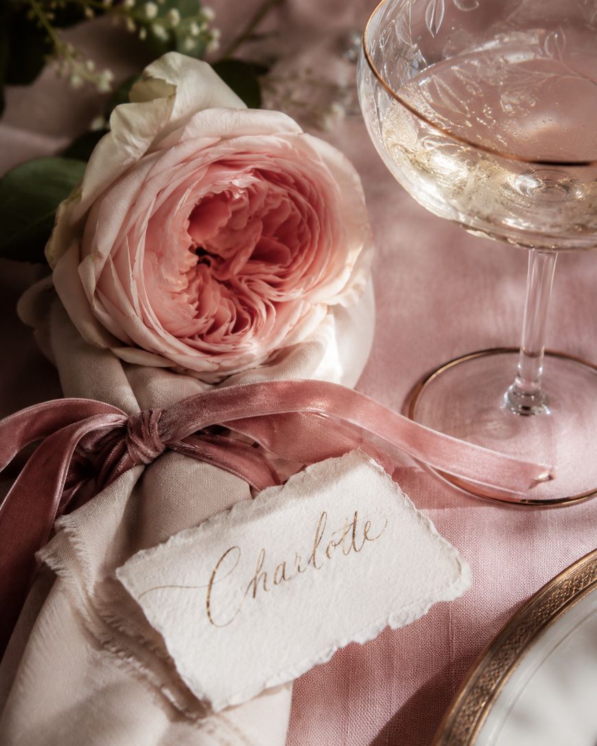 A pink rose and napkin tied with a pink ribbon, a handwritten place card reading Charlotte, and a glass of champagne on a pink tablecloth.