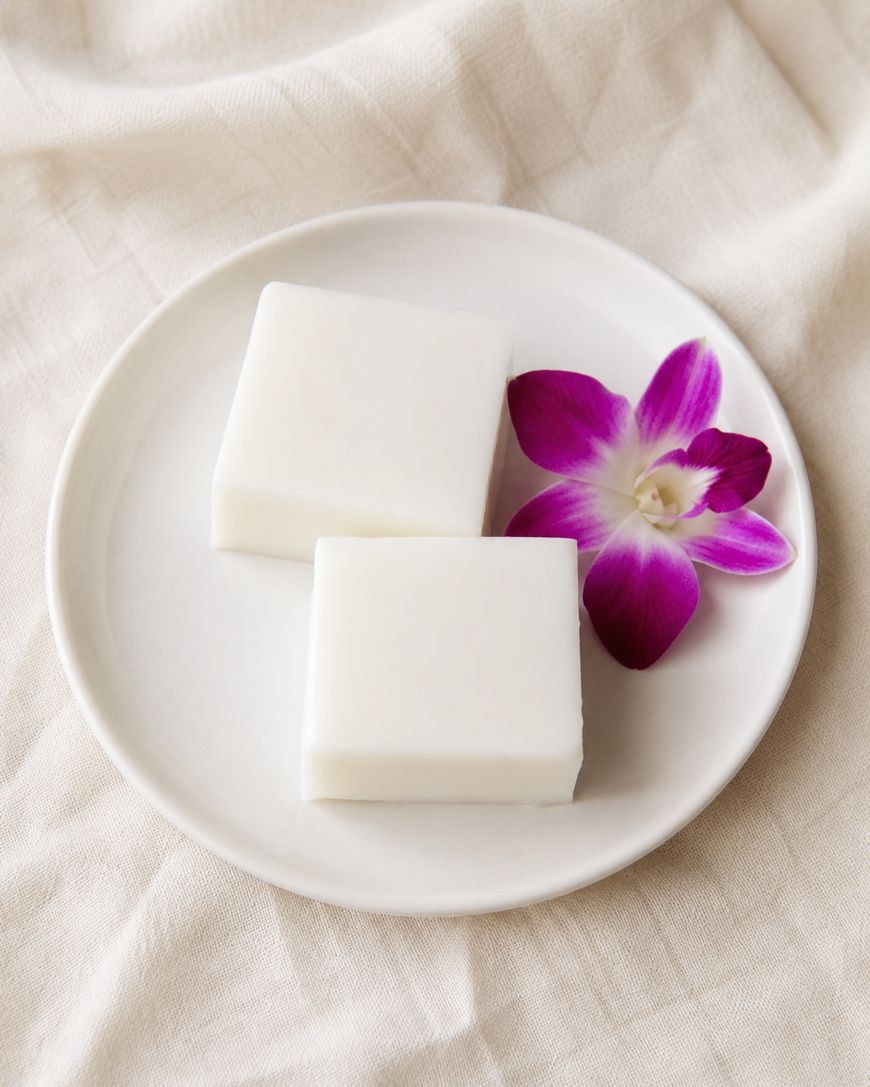 A white plate holds two square pieces of white coconut jelly and a purple orchid flower, set on a cream-colored fabric background.