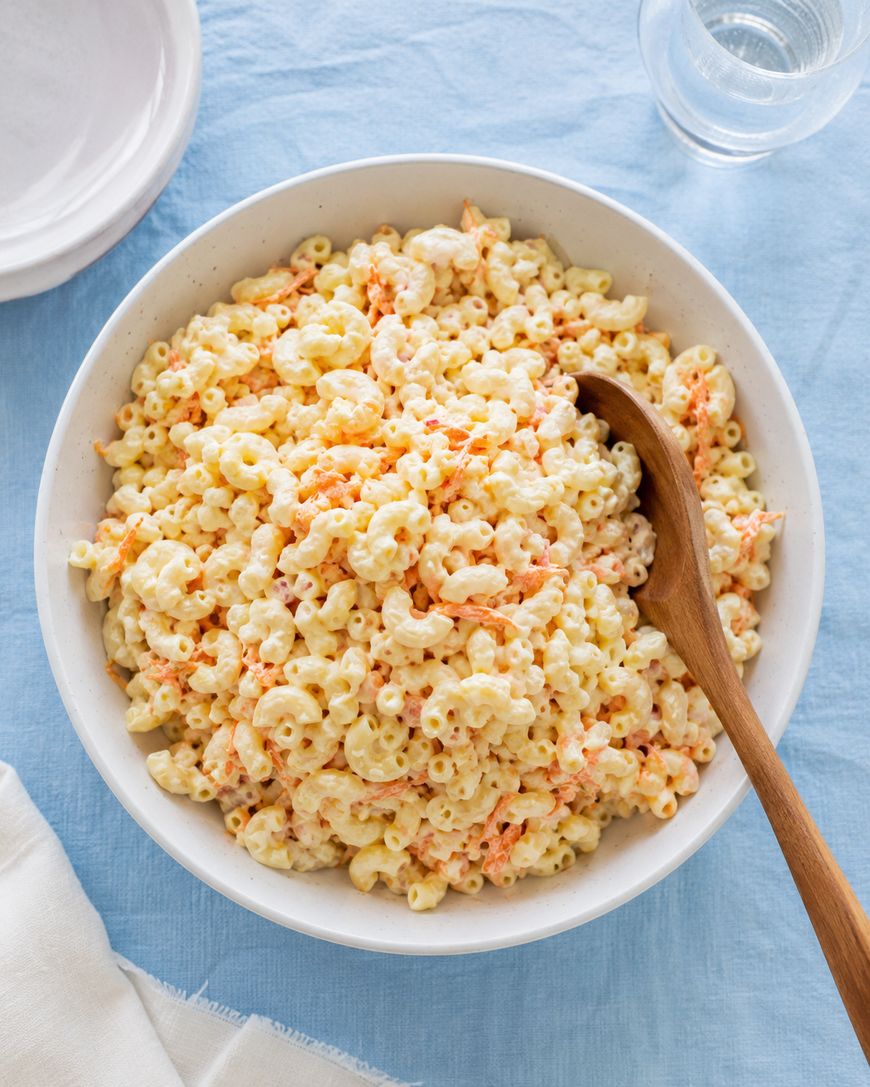 A large white bowl filled with creamy macaroni salad, featuring elbow pasta and shredded carrots, with a wooden spoon resting inside. The bowl is on a light blue tablecloth next to a glass of water and a stack of white plates.