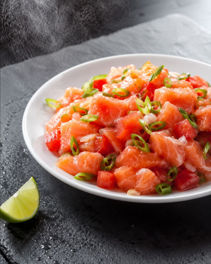 A white plate filled with fresh salmon poke topped with chopped green onions and red bell peppers, placed on a dark slate surface next to a lime wedge.
