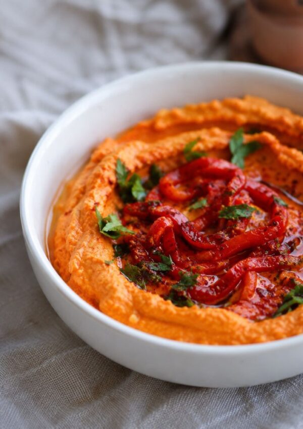 A white bowl filled with creamy red pepper hummus, one of the best healthy dips, garnished with sliced roasted red peppers, olive oil, chopped herbs, and spices, sits on a light gray cloth.