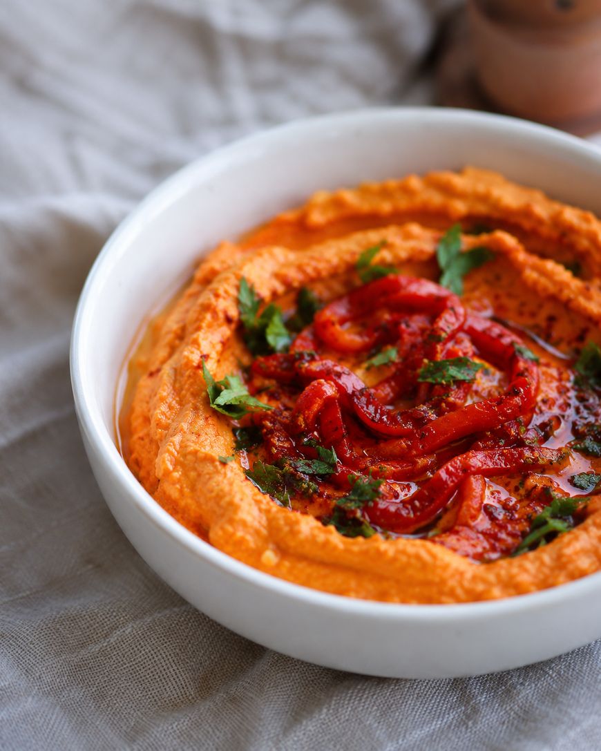 A white bowl filled with creamy red pepper hummus, garnished with sliced roasted red peppers, chopped parsley, olive oil, and spices, set on a light cloth background.