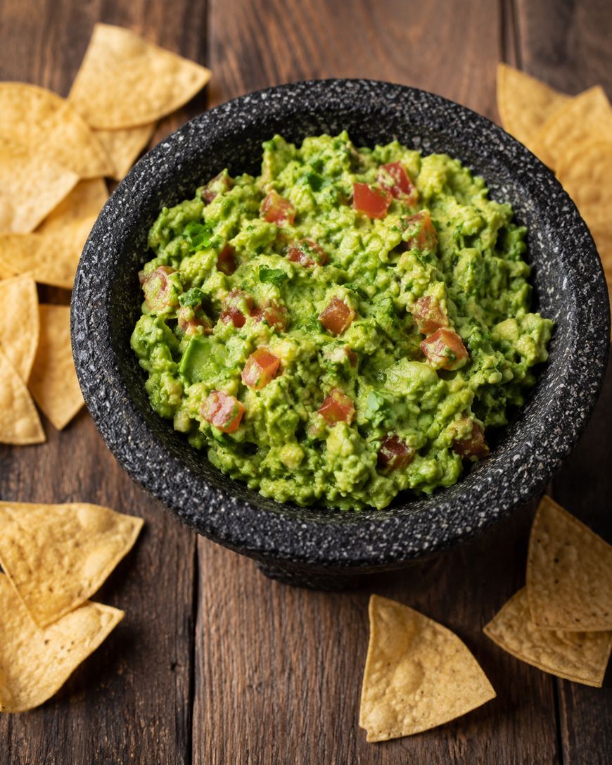 A stone bowl filled with chunky guacamole, garnished with diced tomatoes, surrounded by tortilla chips on a rustic wooden table.