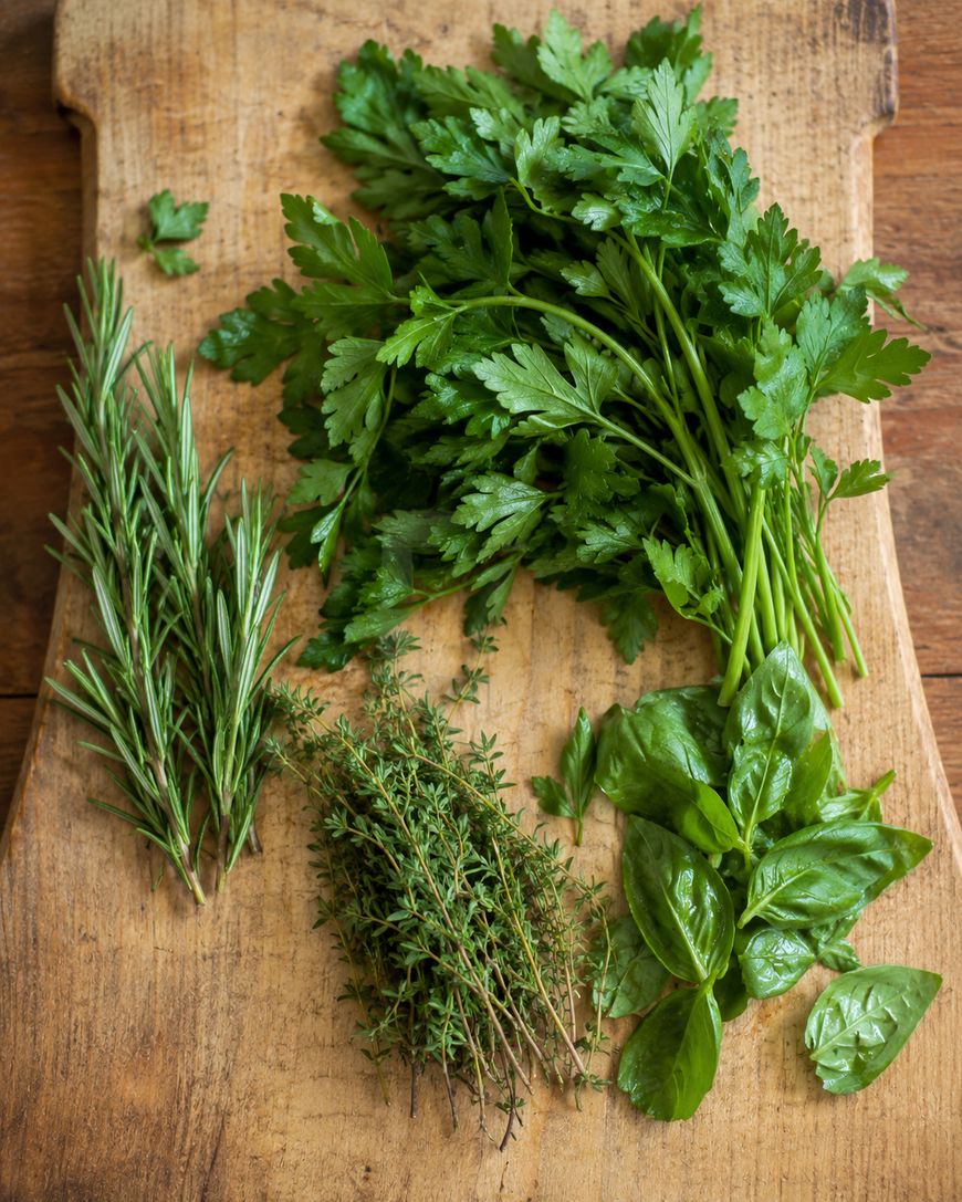 A wooden cutting board with fresh herbs: a bunch of parsley, a cluster of basil leaves, sprigs of rosemary, and sprigs of thyme, all neatly arranged on the board.