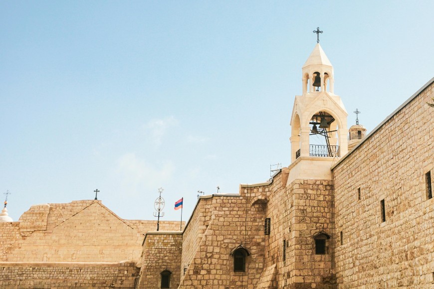 A historic stone church with a bell tower and cross rises against a clear blue sky. An Armenian flag and other crosses are visible on the roof. The architecture features light-colored, textured stone walls.