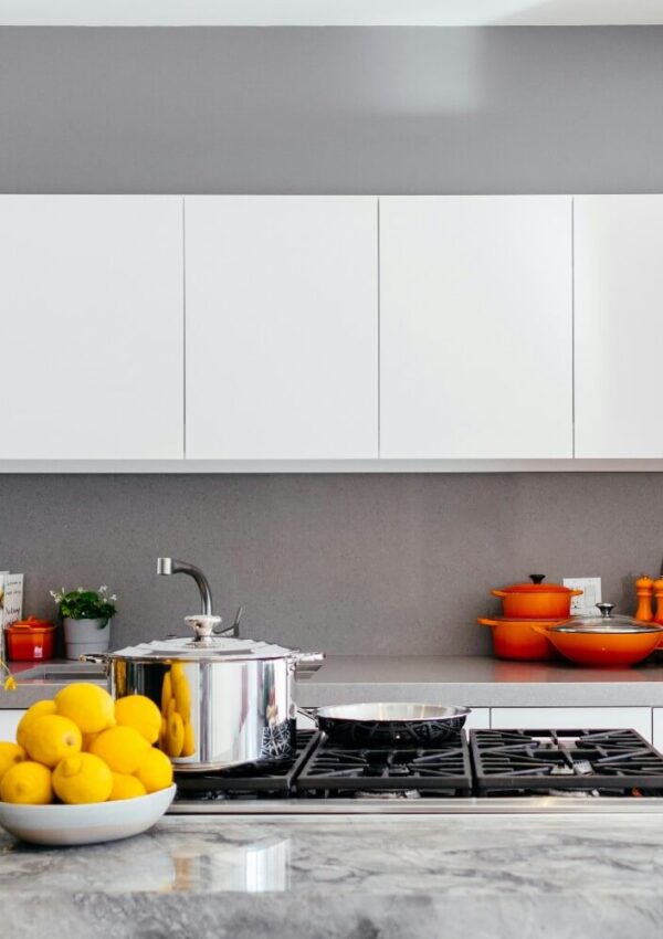 Modern kitchen with white cabinets, a gray backsplash, and a marble countertop. A bowl of lemons, a pot, and a pan are on the stove, with orange cookware and utensils arranged on the counter in the background.