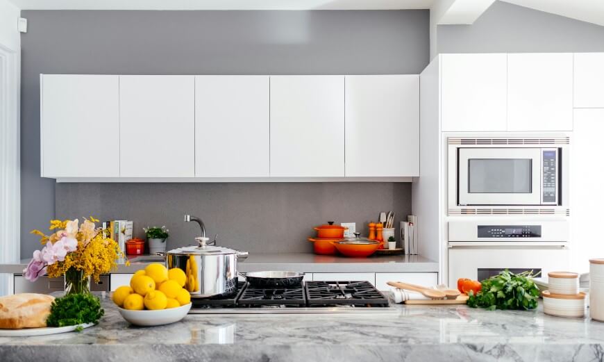 A modern kitchen with white cabinets, a gray backsplash, and a marble countertop. Fresh lemons, herbs, and kitchenware are on the island, and orange pots and a microwave are visible in the background.