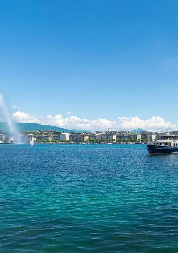A large lake with deep blue water, a fountain spraying high on the left, a boat on the right, and a city skyline with buildings and green hills under a bright blue sky with scattered clouds.