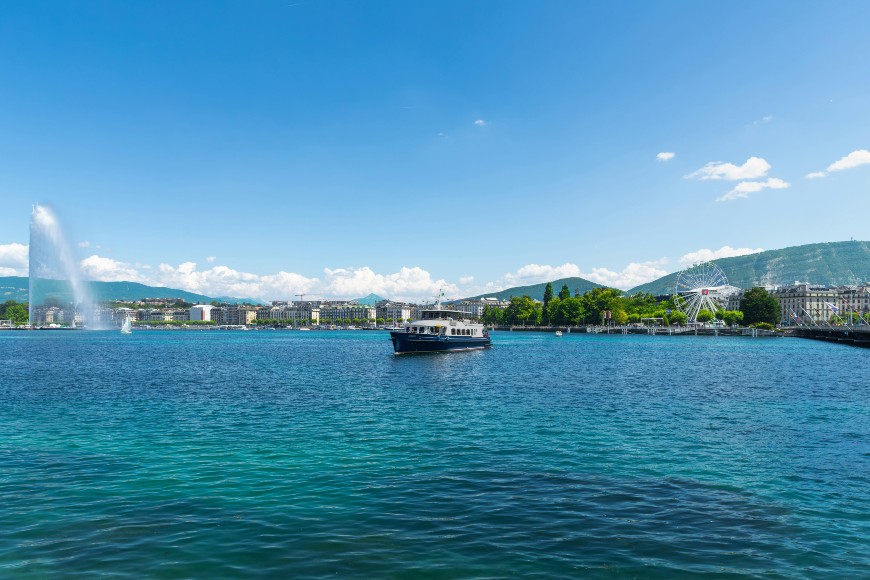 A boat sails on a large blue lake with a fountain spraying water on the left, city buildings, green hills, and a clear sky with scattered clouds in the background.