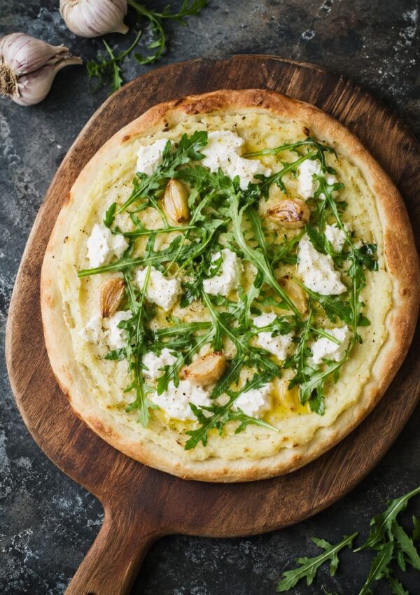 A freshly baked white pizza topped with arugula, roasted garlic cloves, ricotta cheese, and a sprinkling of leftover mashed potatoes—an inventive twist for those seeking creative leftover mashed potato ideas. Served on a wooden pizza peel with fresh toppings.