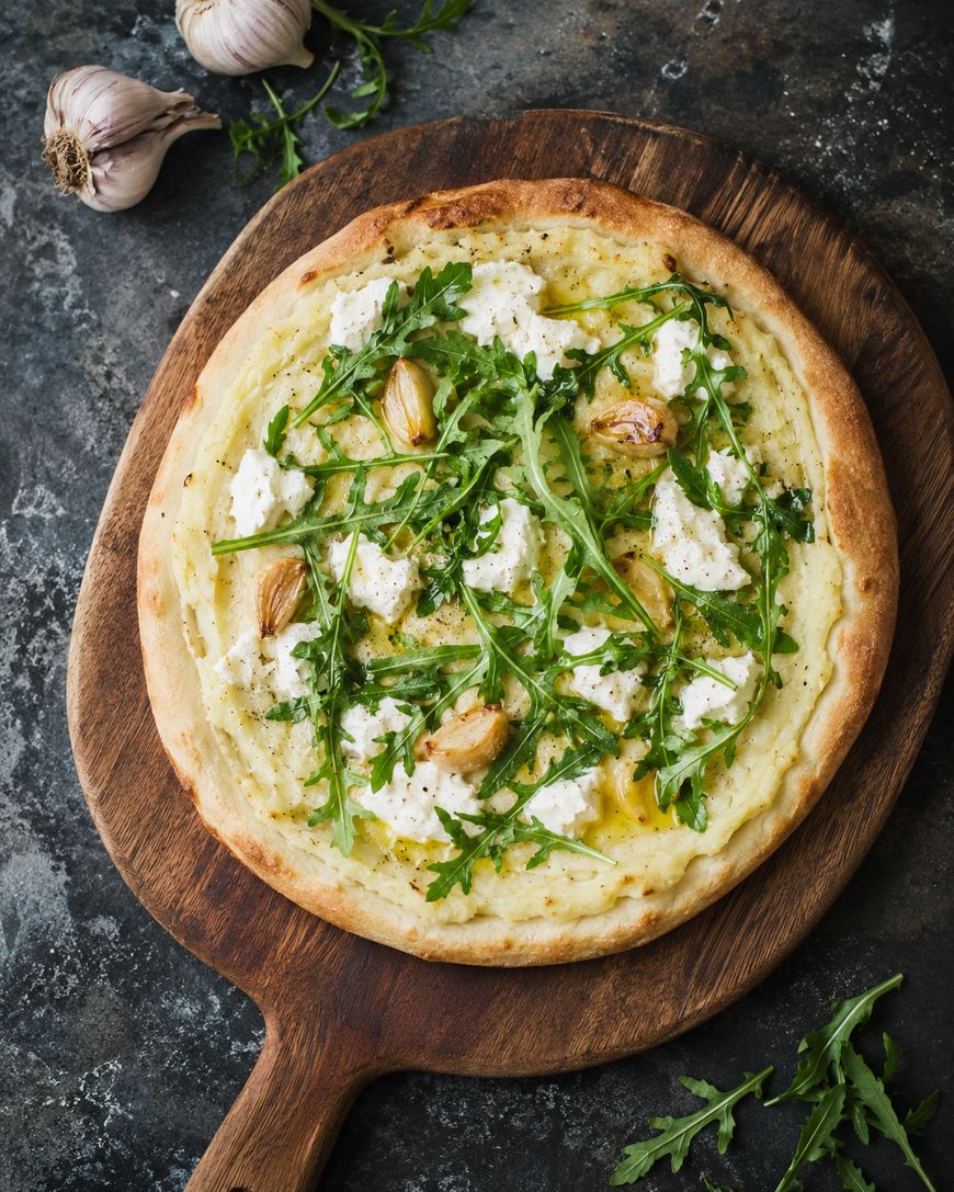 A white pizza topped with ricotta cheese, arugula leaves, and roasted garlic sits on a wooden board. Garlic bulbs and arugula are nearby on a dark stone surface.