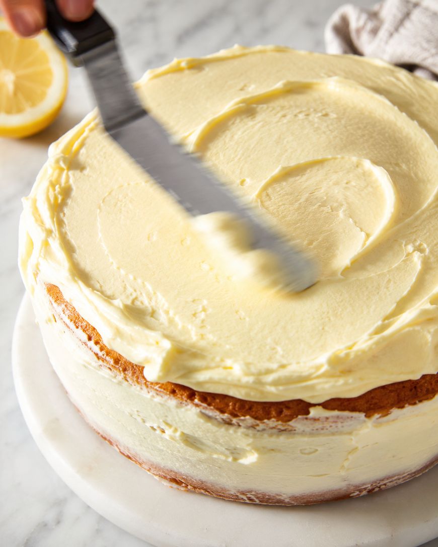 A hand smooths creamy yellow frosting on a round cake using an offset spatula. The cake sits on a marble surface, with a lemon half and a cloth visible in the background.