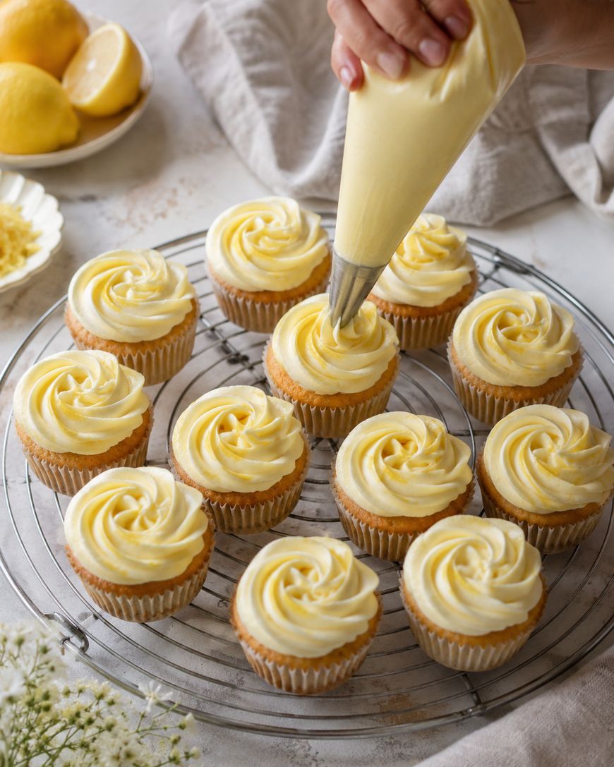 A hand pipes swirls of yellow frosting onto cupcakes arranged on a wire rack, with lemons and a plate of zest in the background.