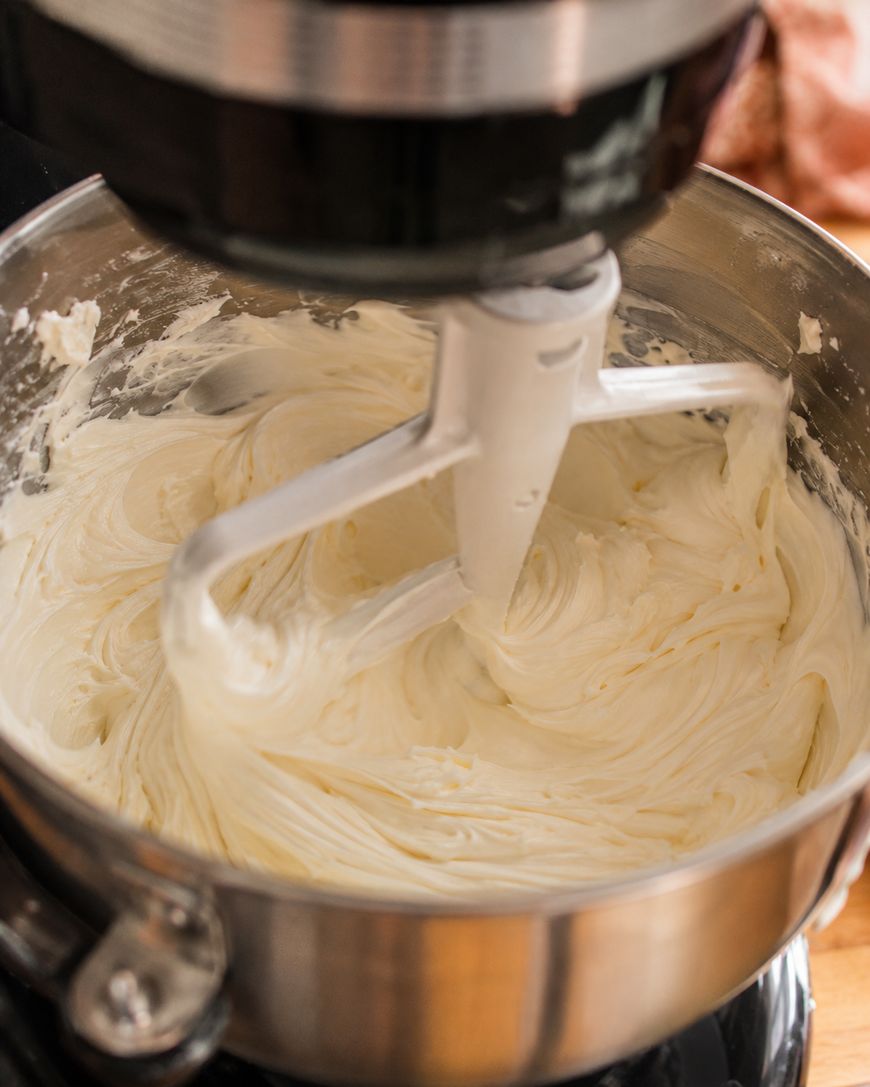 A stand mixer with a paddle attachment is mixing creamy, whipped frosting in a stainless steel bowl. The frosting appears smooth and fluffy, sticking to the sides of the bowl and the paddle.