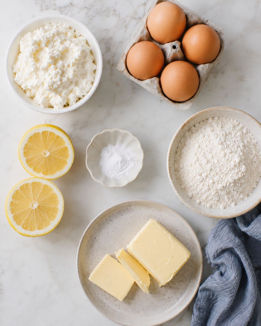A top-down view of baking ingredients on a white surface: a bowl of ricotta cheese, four brown eggs in a carton, two lemon halves, a small bowl of salt, a bowl of flour, and a plate with two butter slices.