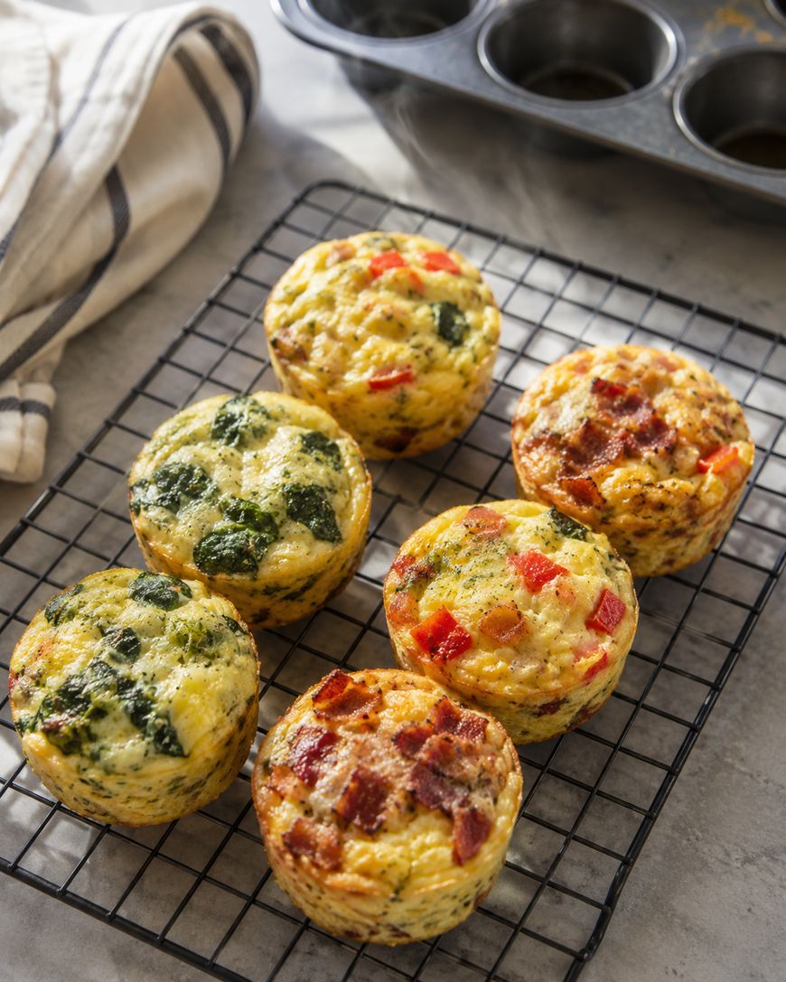 Six baked egg muffins with various fillings like spinach, bacon, and red peppers sit on a cooling rack. A striped towel and a muffin tin are nearby on a light countertop.