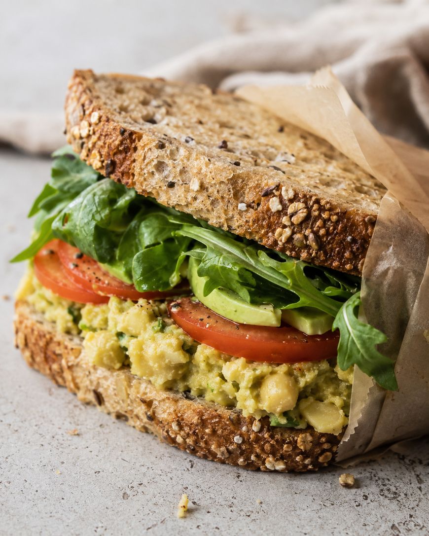A sandwich made with multigrain bread, filled with mashed chickpeas, avocado slices, tomato slices, and fresh arugula, partially wrapped in brown parchment paper, sits on a light surface.