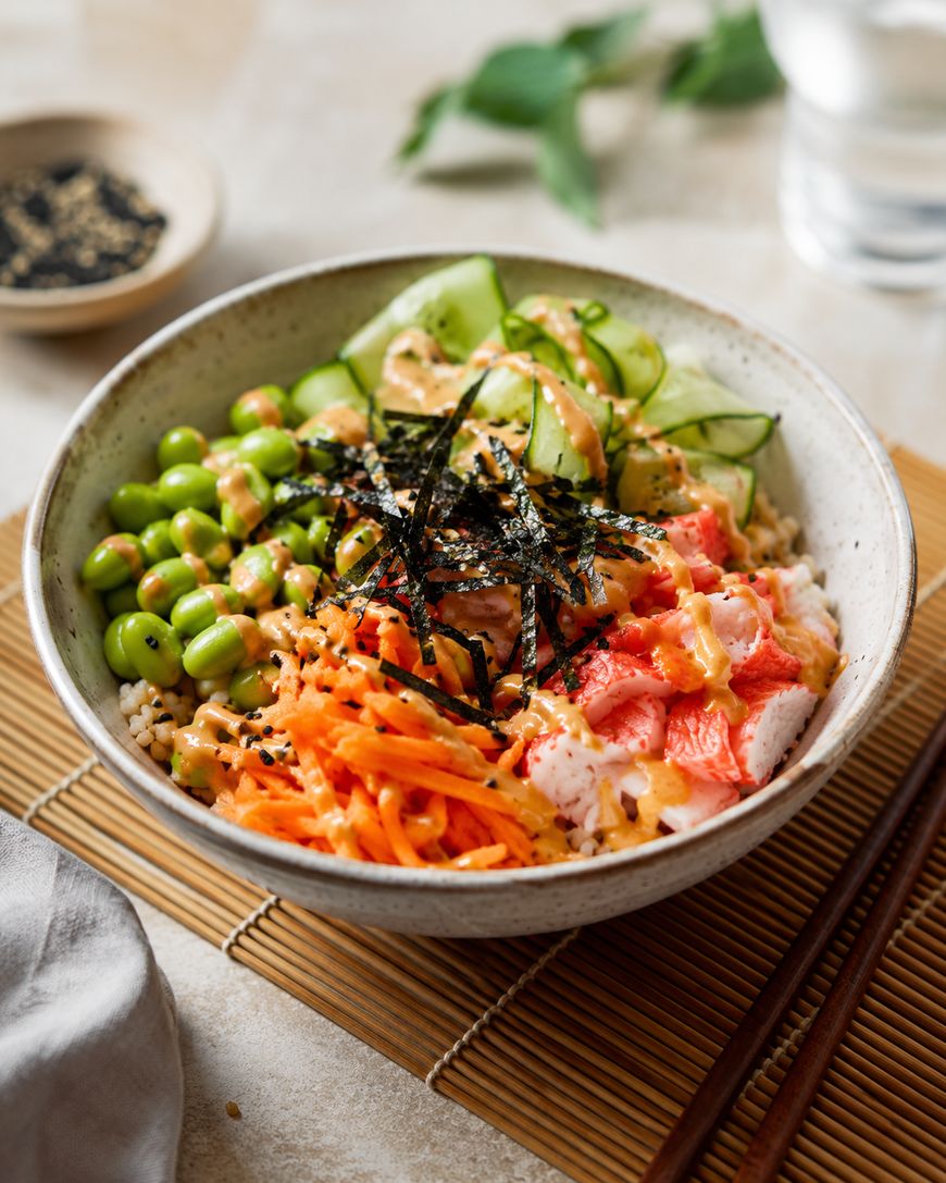 A poke bowl with white rice, imitation crab, shredded carrots, edamame, cucumber slices, seaweed strips, sesame seeds, and creamy sauce, served in a ceramic bowl with chopsticks on a bamboo mat.