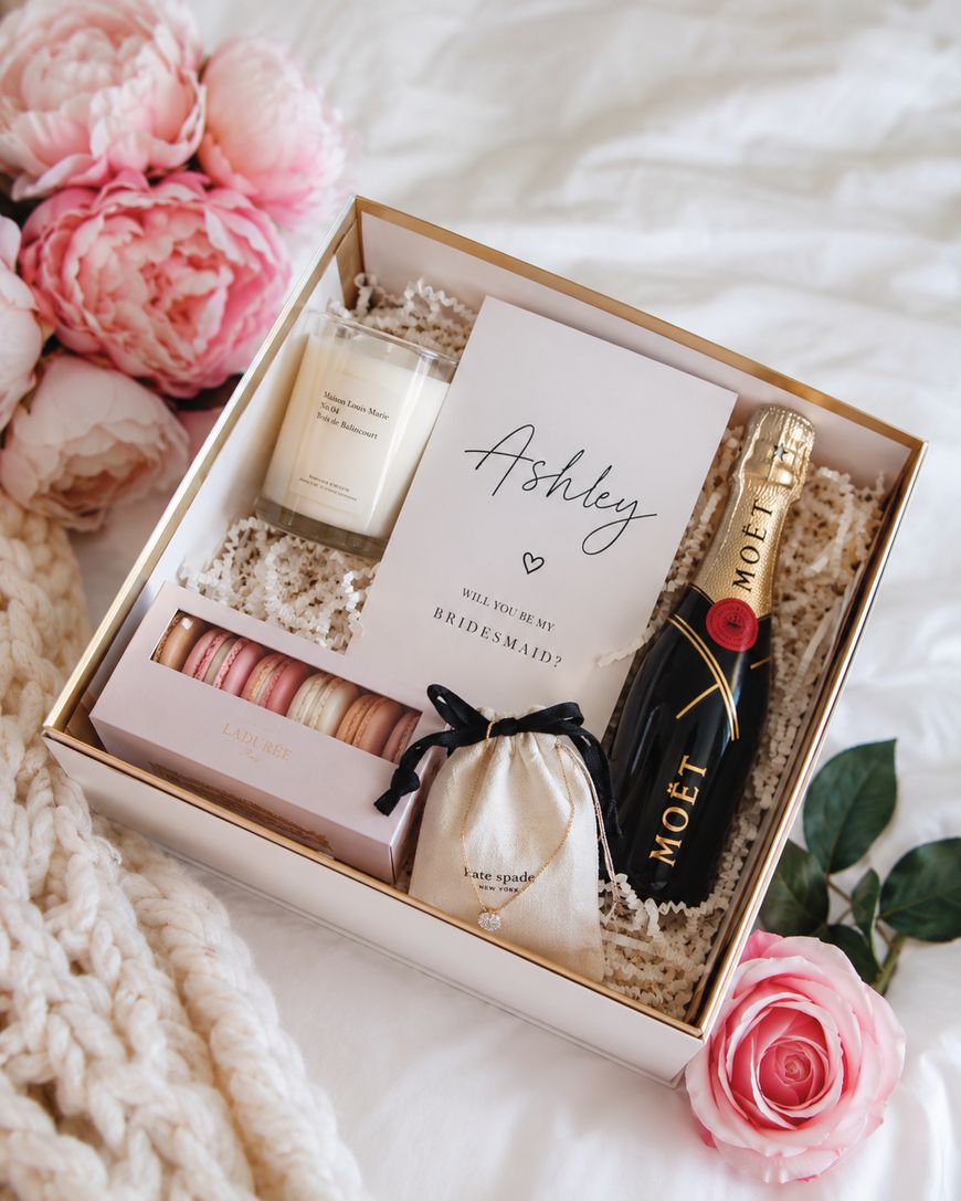 A bridesmaid proposal gift box containing a candle, macarons, a gold bag with jewelry, a Will you be my bridesmaid? card, and a bottle of Moët champagne, surrounded by pink flowers and a cozy knit blanket.