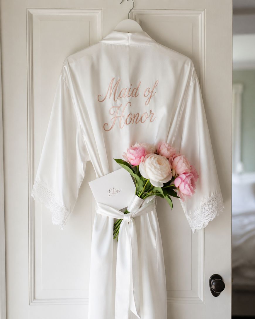 A white satin robe with Maid of Honor embroidered in pink hangs on a door. A bouquet of pink and white flowers and an envelope labeled Elise are tucked into the robes sash.