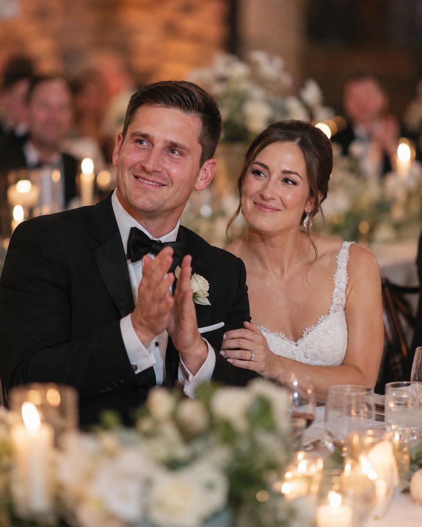 A bride and groom, dressed in wedding attire, sit closely together at a candlelit reception table, smiling and clapping as they enjoy a special moment during their celebration.