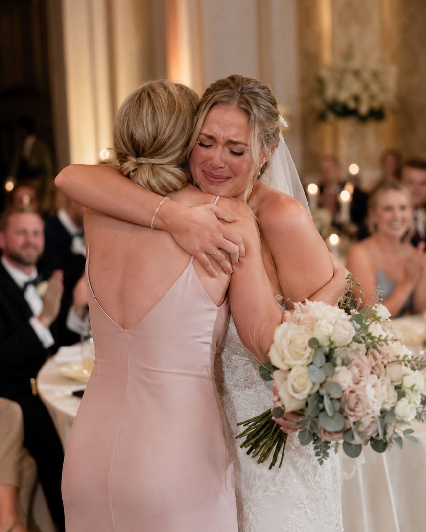 A bride in a wedding dress and veil hugs a woman in a light pink dress, both emotional. The bride holds a bouquet of white and blush flowers. Guests seated at tables in formal attire are visible in the background.