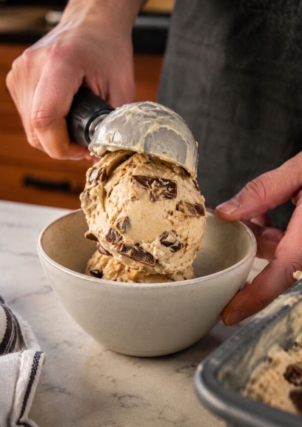 A person scoops two large scoops of chunky chocolate chip homemade ice cream into a bowl on a marble countertop, with a striped towel and an ice cream container nearby.