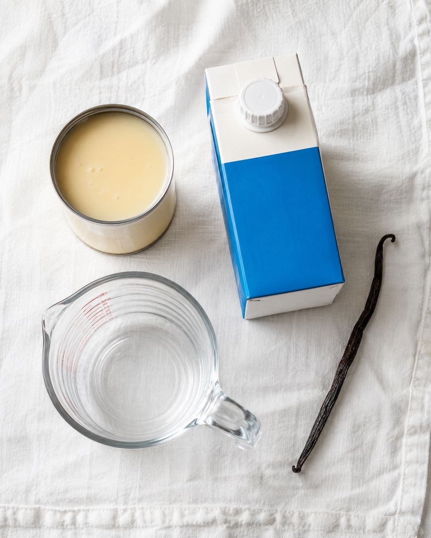 A can of condensed milk, a blue and white milk carton, an empty glass measuring cup, and a vanilla bean on a white cloth background.