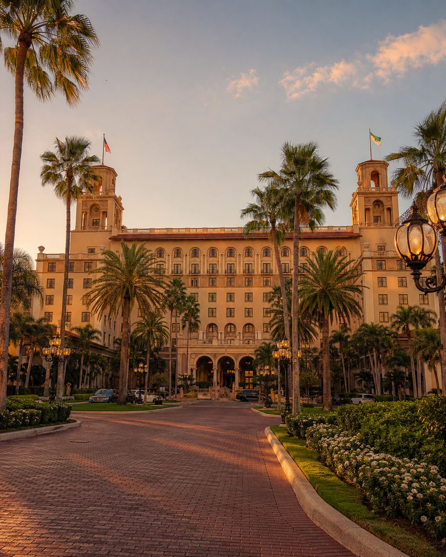 A grand hotel with two towers, palm trees, and flags on the roof is bathed in golden sunlight. The building is framed by lush landscaping and a wide brick driveway.