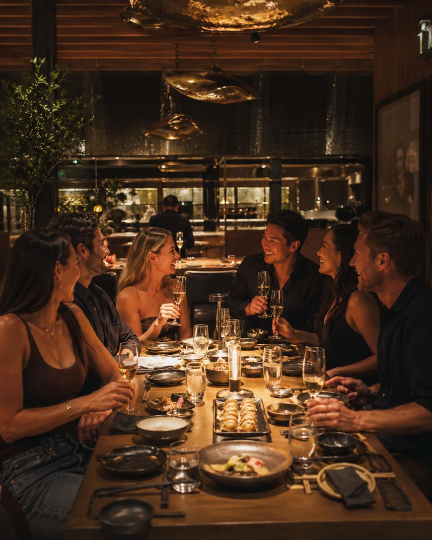 Six people sit around a warmly lit restaurant table, smiling and raising glasses in a toast. Plates of food and drinks are spread across the table, creating a lively and celebratory atmosphere.