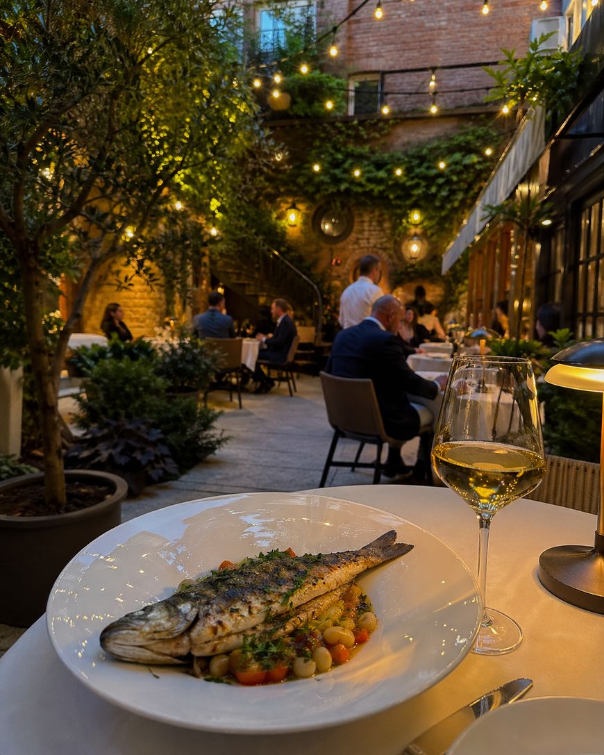 A grilled whole fish on a plate with vegetables is set on a table with a glass of white wine in a cozy, elegant outdoor restaurant with string lights, plants, and diners in the background.